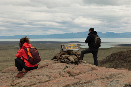 Pai e filha no Mirador de las Águilas, na Patagônia, com mochilas Allca Allcatrazes: ela agachada fotografando e ele em pé diante do lago e da cordilheira