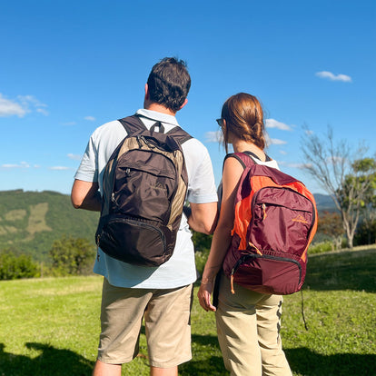 Mochila Charlie Dobrável 25L Allca Allcatrazes em nylon impermeável - casal em trilha ao ar livre usando as versões Café com Areia e Bordô com Laranja nas costas