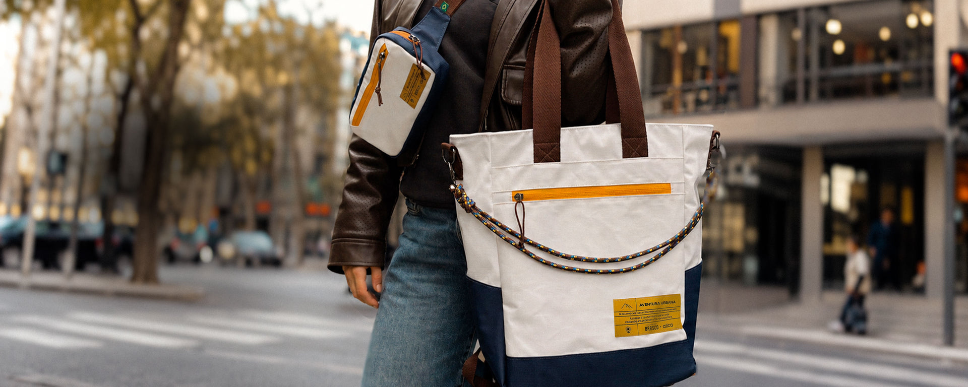 Pessoa vestindo jaqueta marrom-escura e calça jeans azul em uma rua urbana com calçamento de pedras. Ela usa uma pochete transversal branca e azul-marinho com zíper laranja e etiqueta em couro, além de carregar uma tote bag branca e azul-marinho da marca allcatrazes é uma collab da allca com o Mercado brasco. com alças marrons, cordão estampado na parte frontal, zíper laranja e etiqueta em couro. Ao fundo, árvores e prédios aparecem desfocados sob luz natural, criando um ambiente urbano elegante.